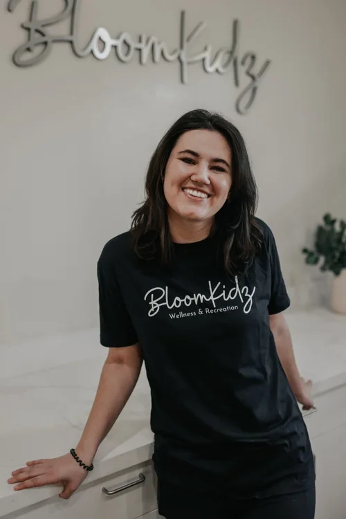 A smiling woman with dark hair, wearing a black BloomKidz Wellness & Recreation t-shirt, standing in front of a white wall with a sign that reads "BloomKidz" and a potted plant in the background.
