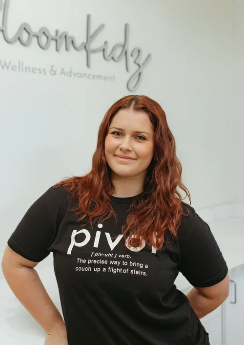A woman with long, wavy red hair standing in front of a white wall with the word 'wellness' in cursive. She is smiling, wearing a black t-shirt with the word 'pivotal' and its definition printed on it.