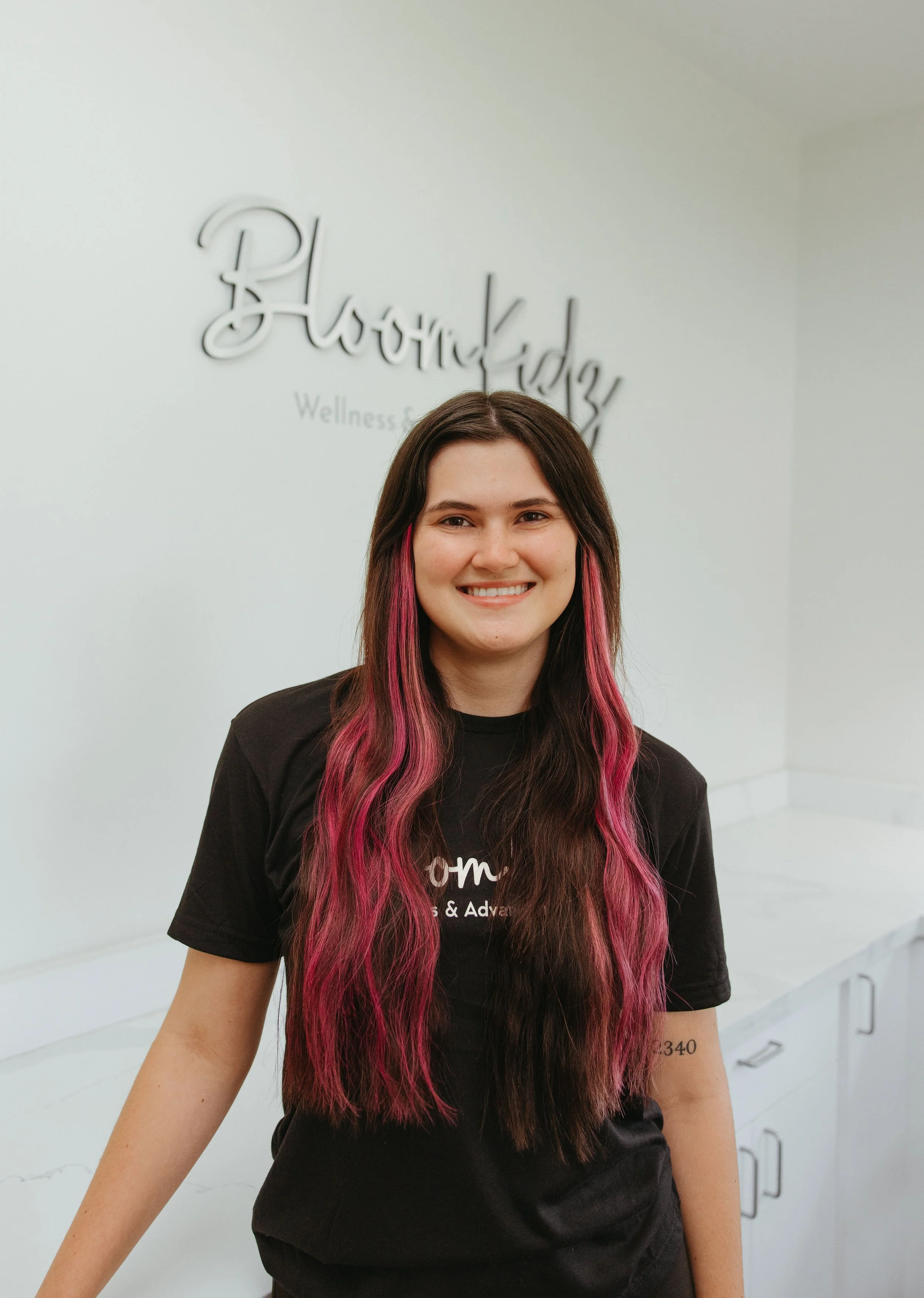 A woman with brown hair and pink highlights smiling at the camera, wearing a black T-shirt, in a clinic with a white wall and a sign that reads 'BloomKidz'.