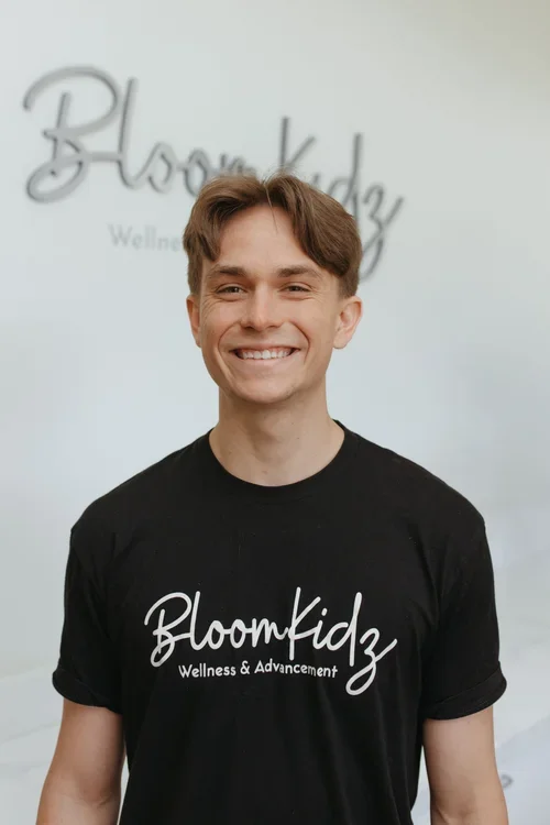 Young man smiling, wearing a black shirt with the logo 'BloomKidz Wellness & Advancement,' standing in front of a white wall with a similar logo in the background.
