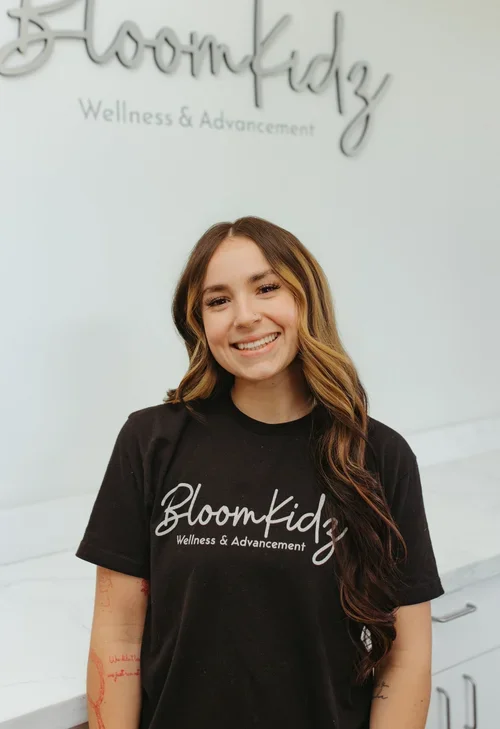 A young woman with long wavy hair smiling at the camera, wearing a black t-shirt with 'Bloomkidz Wellness & Advancement' written on it, standing in front of a white wall with the same logo.