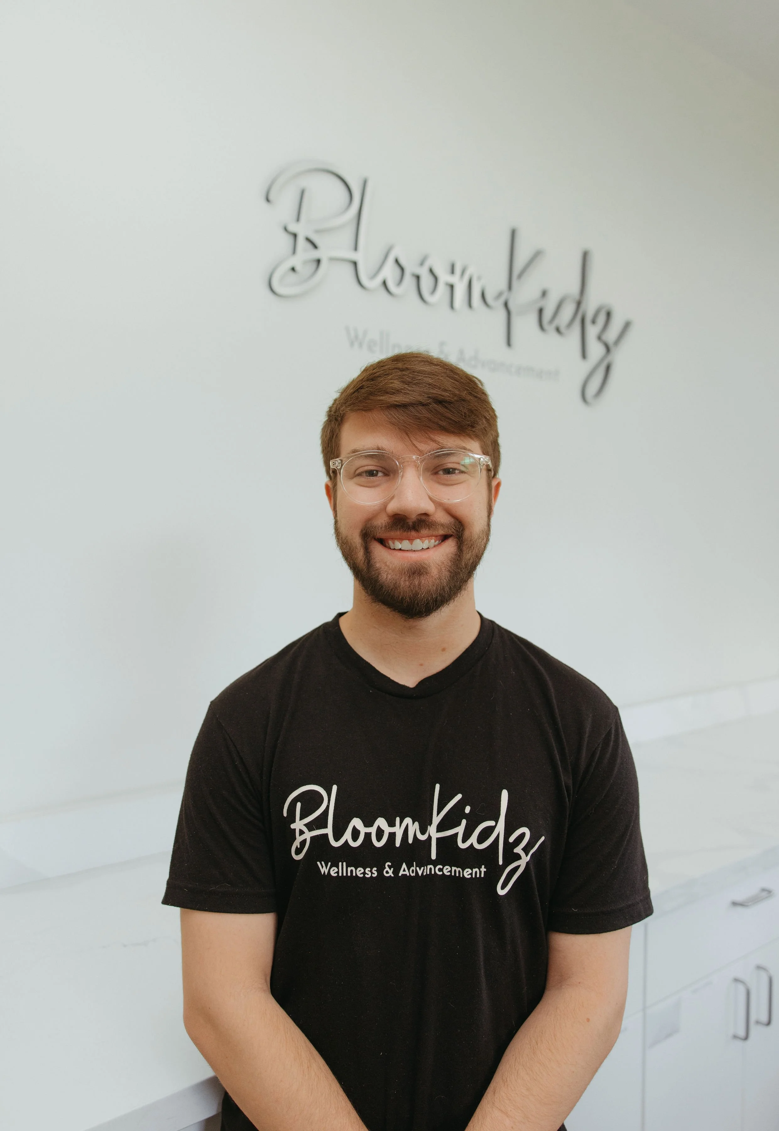 A smiling young man with glasses and a beard standing in front of a white wall with the words 'BloomKidz' and 'Wellness & Advancement' written above him. He is wearing a black T-shirt with similar text.