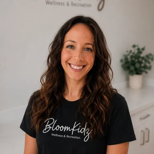 Smiling woman with long wavy brown hair wearing a black Bloom Kidz Wellness & Recreation t-shirt standing indoors near a white cabinet and a potted plant in the background.