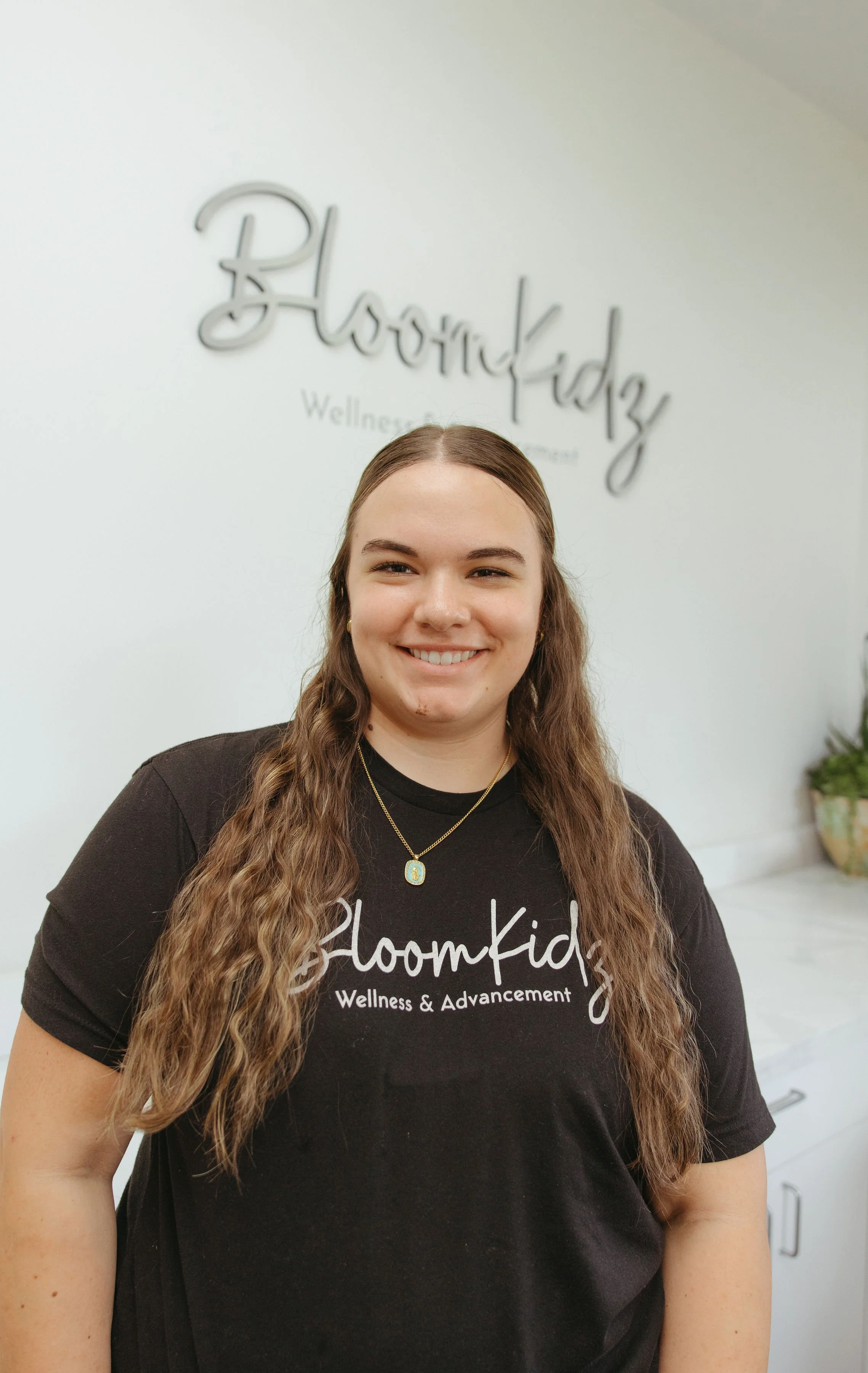 Smiling woman with long brown hair wearing a black BloomKidz Wellness & Advancement T-shirt