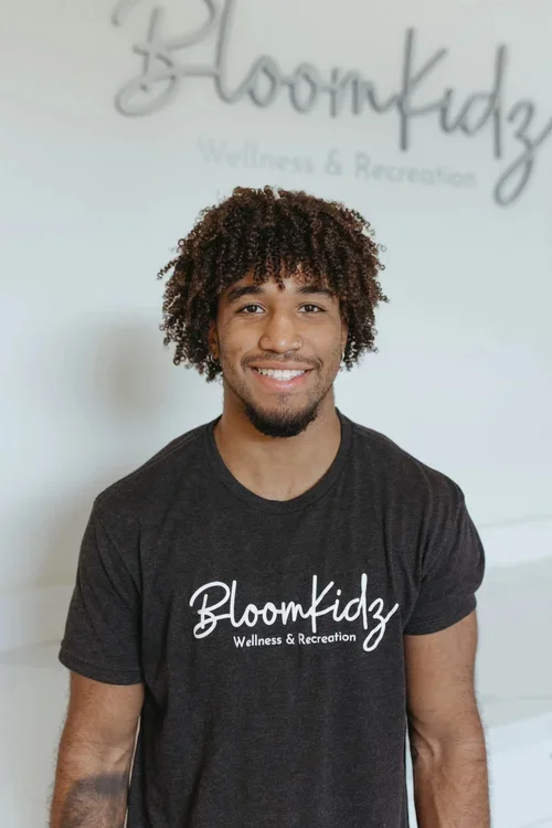 Smiling young man with curly hair and facial hair, wearing a black t-shirt with 'Bloomkidz Wellness & Recreation' logo, standing indoors in front of a white wall with the same logo in the background.