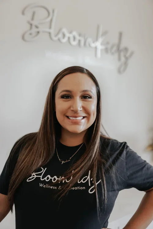 A woman with long brown hair smiling at the camera, wearing a black t-shirt with white text, standing in front of a wall with the word "BloomKidz" written in large cursive.