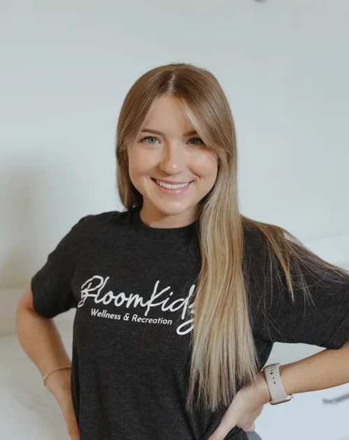 A young woman with long blonde hair smiling and wearing a black t-shirt with white text. She has her hands on her hips and wears a white watch. The background is a light-colored wall.