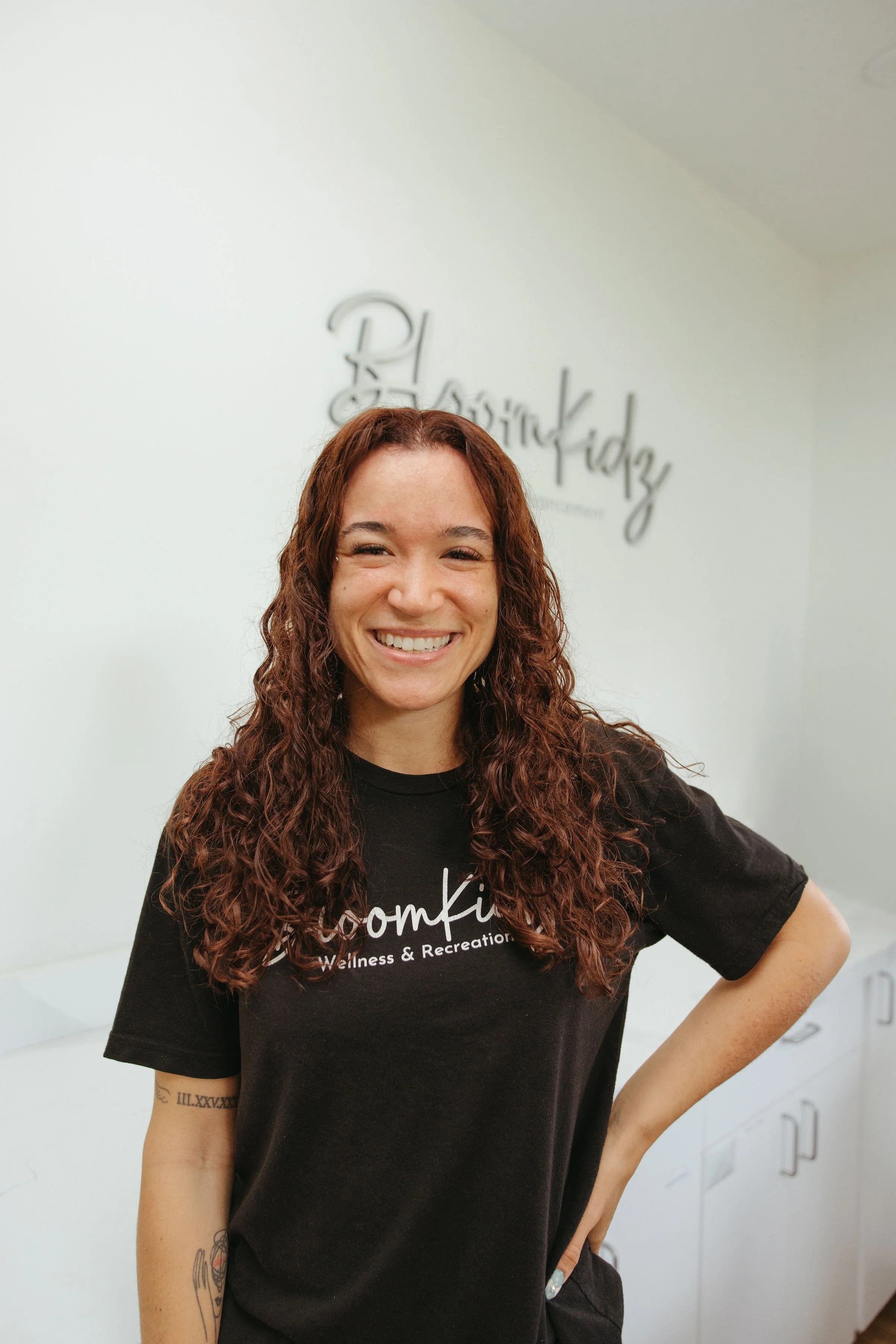 Smiling woman with long curly hair wearing a black wellness and recreation T-shirt in front of a white wall.