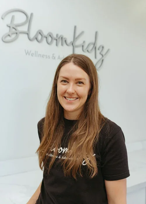 A woman with long brown hair and a black shirt smiling at the camera in front of a light-colored wall with the logo 'Bloomkidz Wellness & Advancement' in the background.