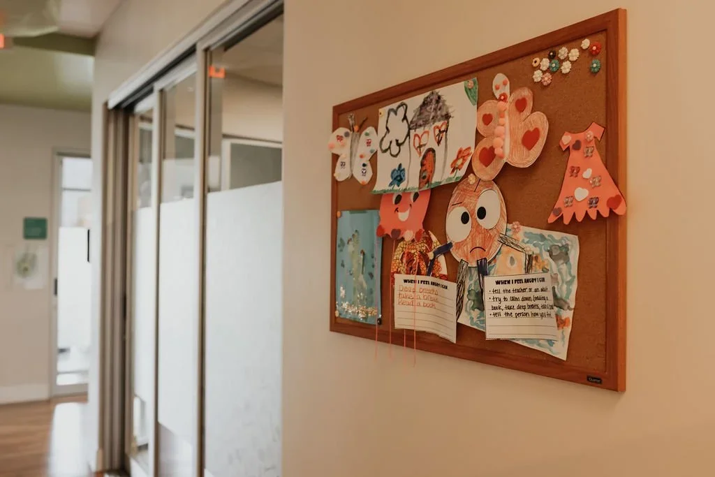 Children's artwork and crafts pinned on a bulletin board in a hallway, including paper flowers, butterflies, and drawings.