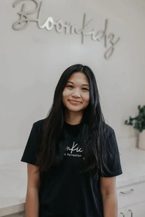 Young woman with long black hair smiling in an indoor space with a sign behind her that says 'BloomKidz' and a potted plant.