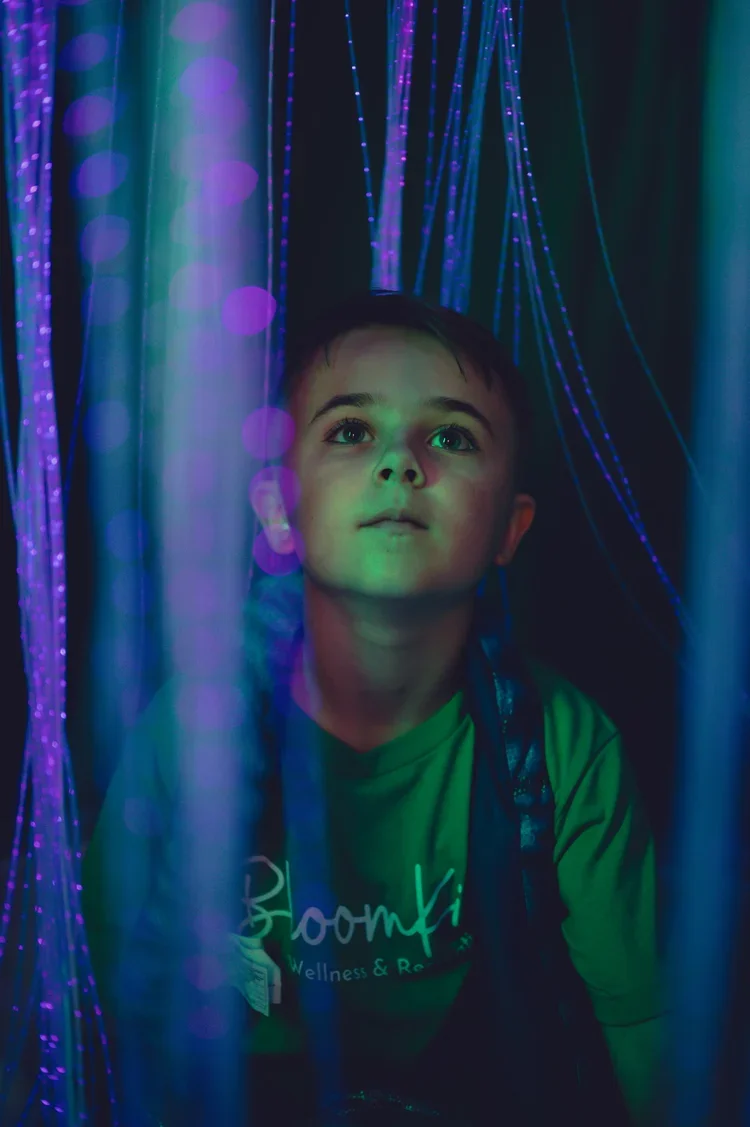Young boy looking up in a dark room illuminated by colorful, glowing lights and strings of blue and purple LED lights.