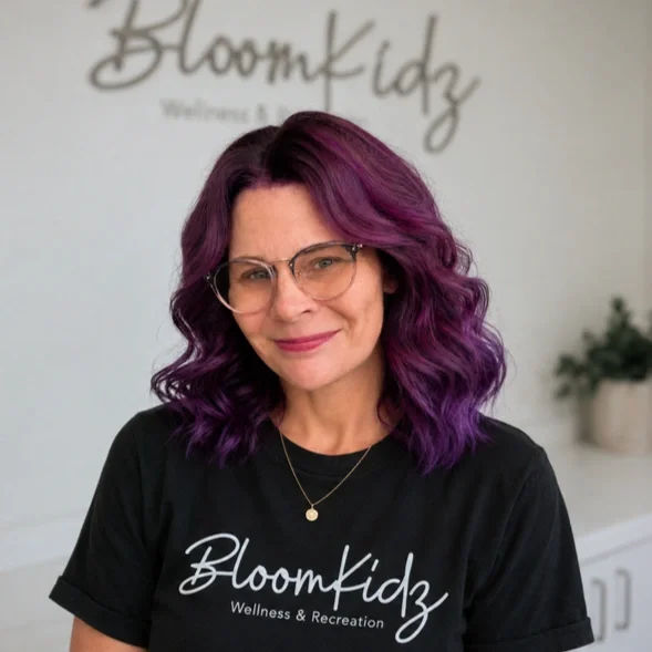 A woman with curly purple hair and glasses smiling in front of a wall with the Bloomkidz logo, wearing a black T-shirt with the same logo.
