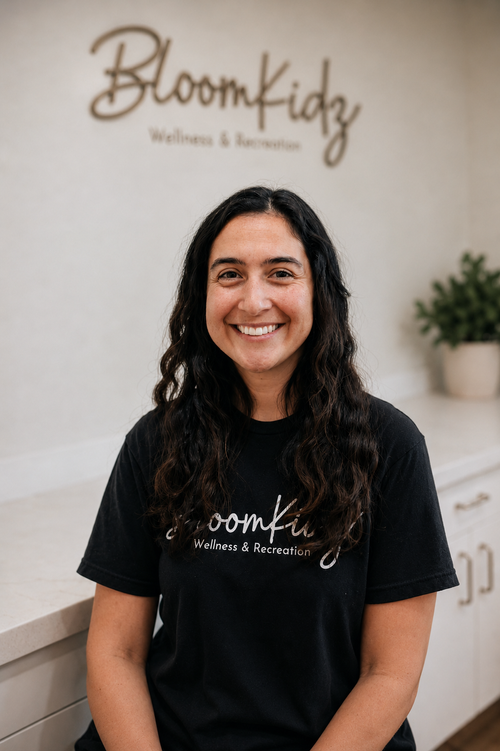 Smiling woman with long dark hair wearing a black Bloomfidz Wellness & Recreation T-shirt inside a wellness center with a sign and potted plant in the background.