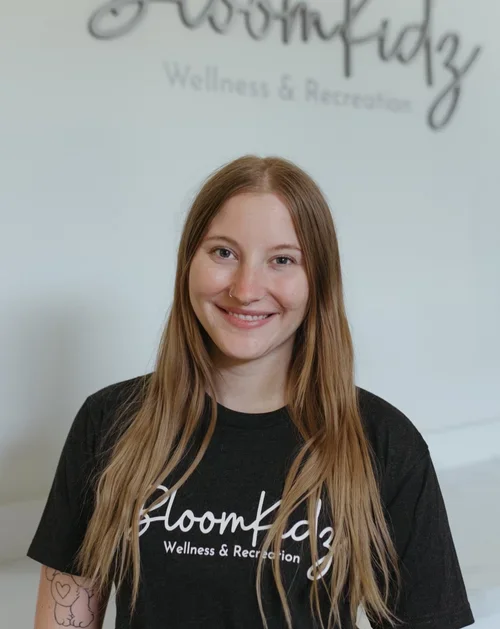 A smiling woman with long, light brown hair stands in front of a wall with a logo that reads 'Bloomkidz Wellness & Recreation'. She is wearing a black T-shirt with the same logo.