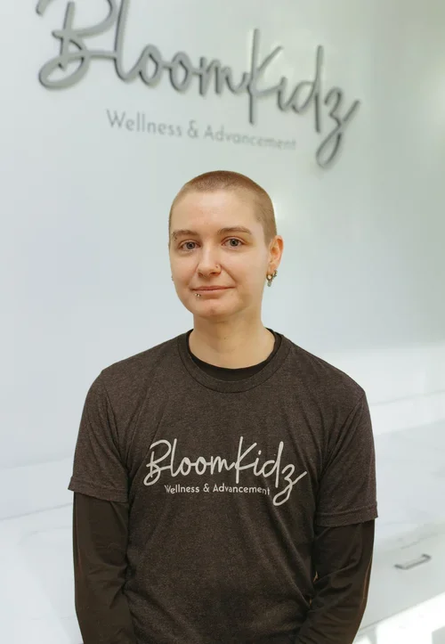 A woman with a shaved head wearing a black BloomKidz Wellness & Advancement t-shirt standing in front of a white wall with the BloomKidz logo.