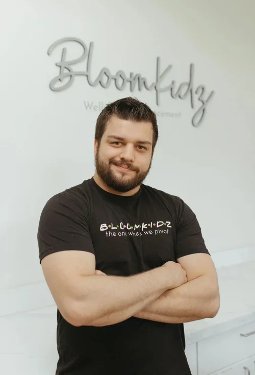 A man with a beard and short hair smiling with crossed arms in front of a BloomKidz sign on a wall.