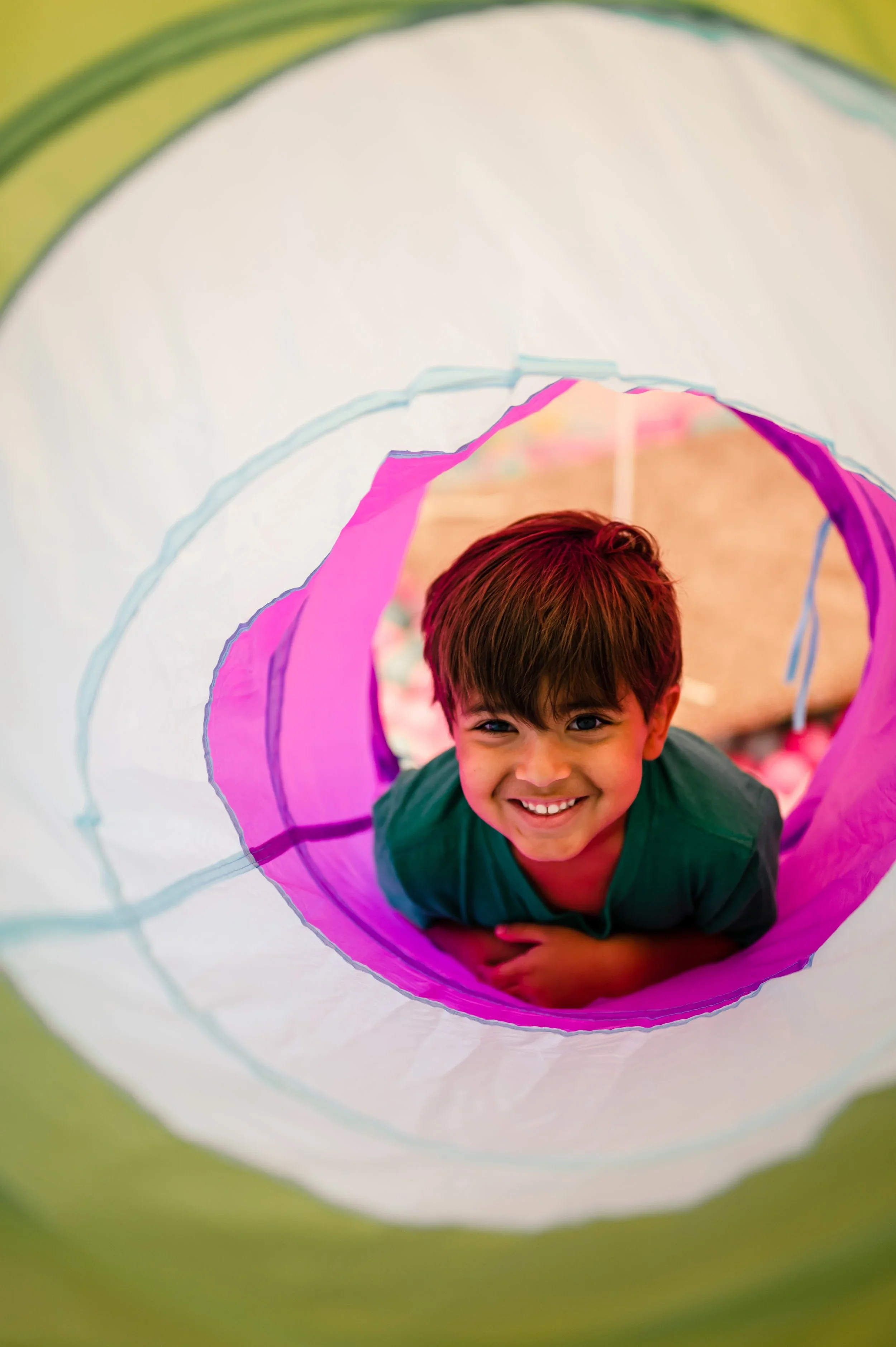 A young boy with brown hair and a big smile crawling through a colorful play tunnel with pink, blue, and white sections.