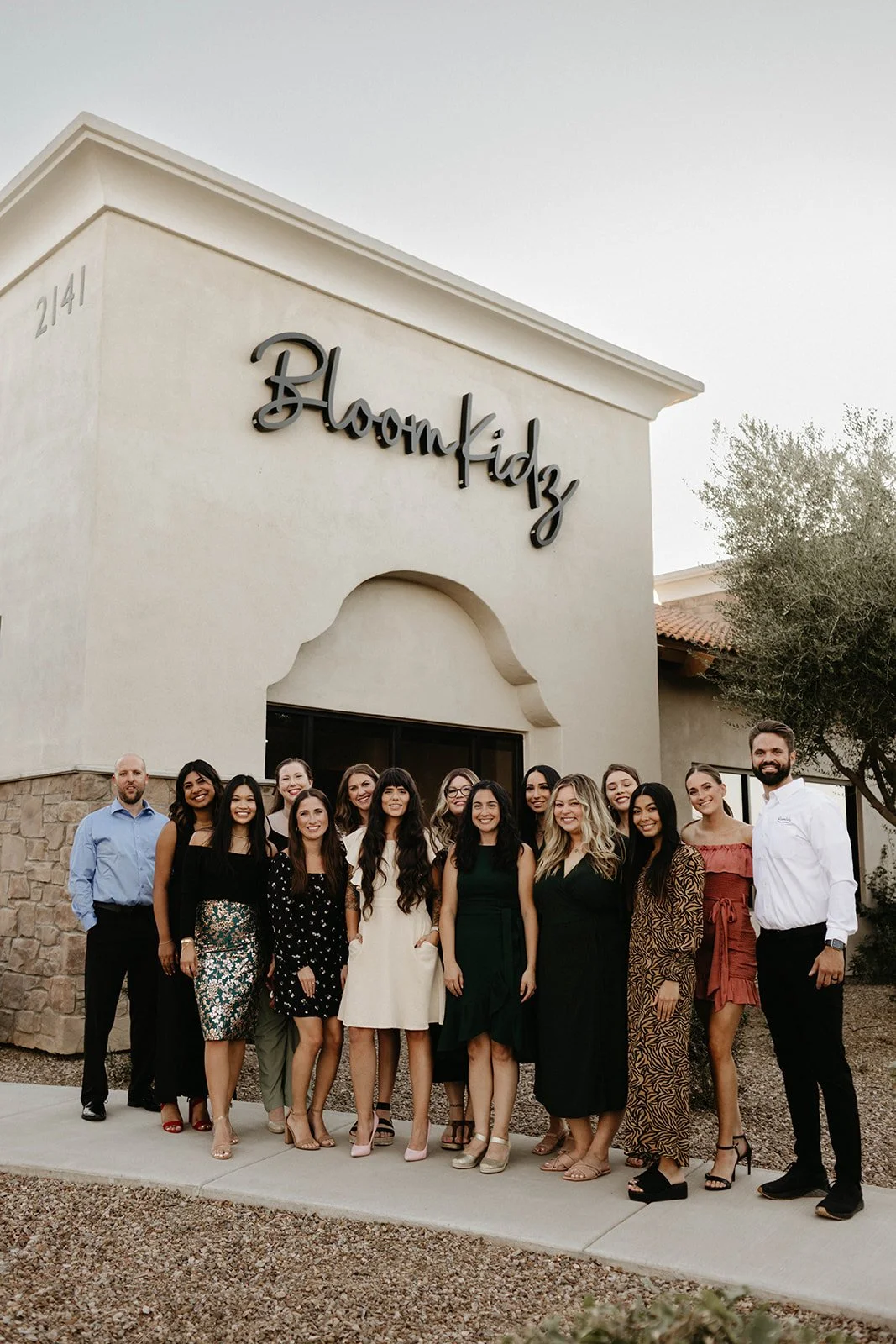 A group of fifteen people, mostly women, standing in front of a building with a sign that reads 'BloomKidz'. They are dressed in semi-formal attire, smiling for the photo.