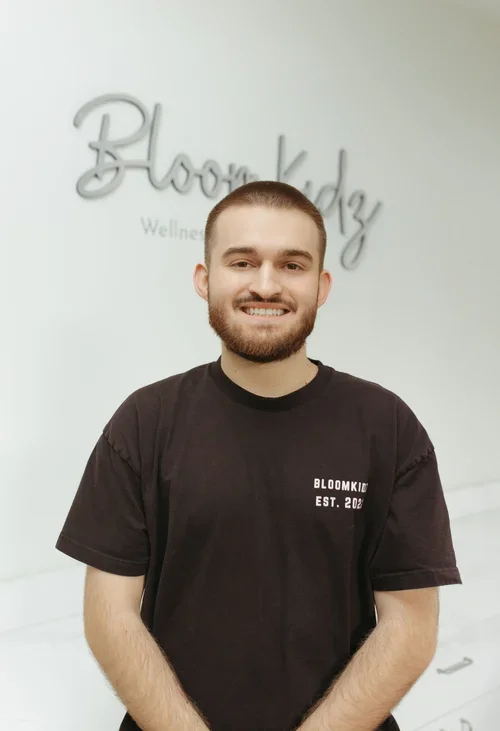 A smiling man with a beard and short hair standing in front of a wall with 'Bloom Kidz' and 'Wellness' written on it, wearing a black t-shirt with 'BLOOMKIDZ EST. 2022' printed on it.