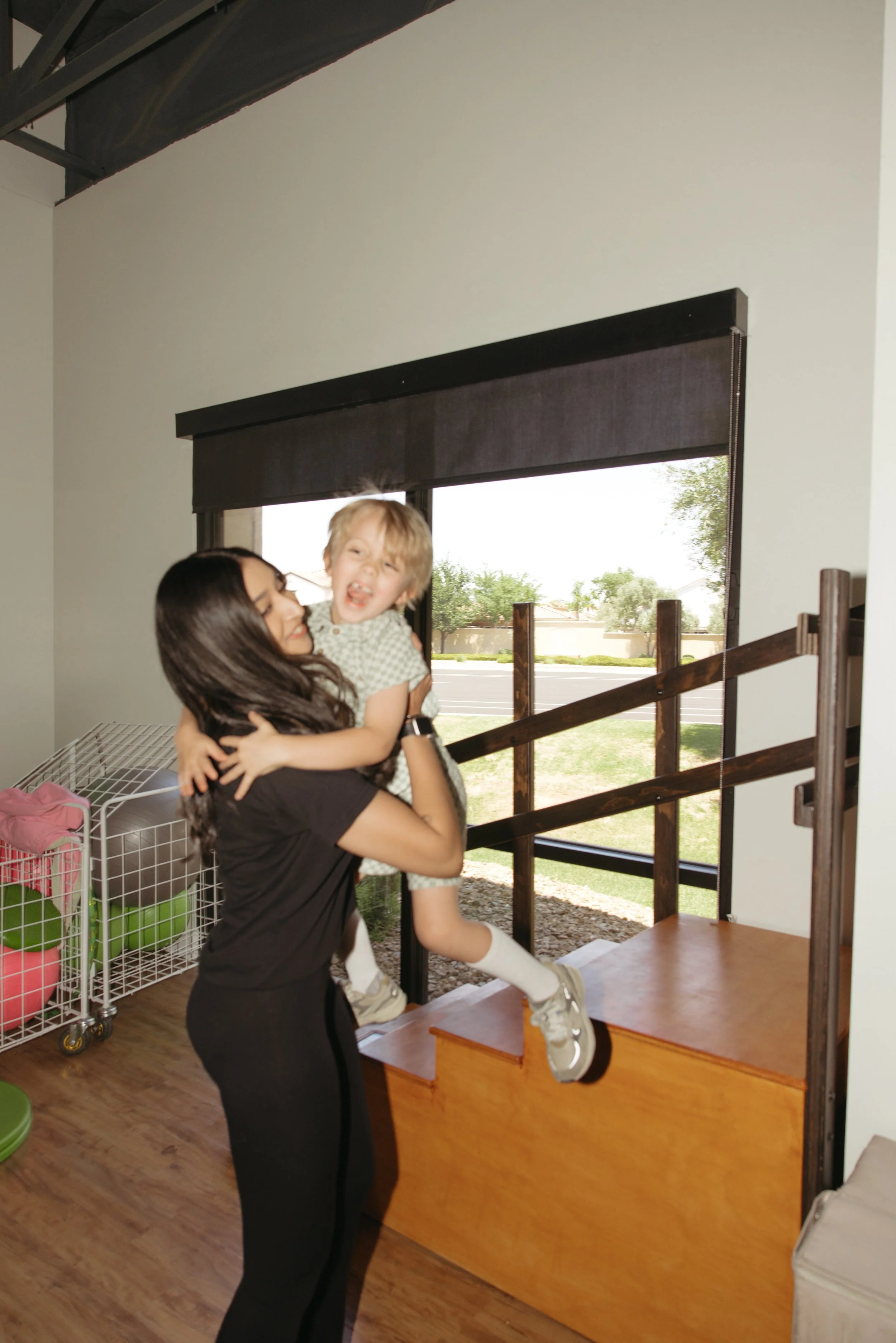 A woman lifting a young child in the air inside a room with large windows and a wooden staircase railing.
