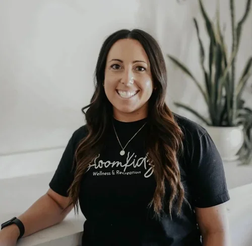 A smiling woman with long dark hair wearing a black t-shirt with white text and a necklace, standing indoors with a large plant in the background.
