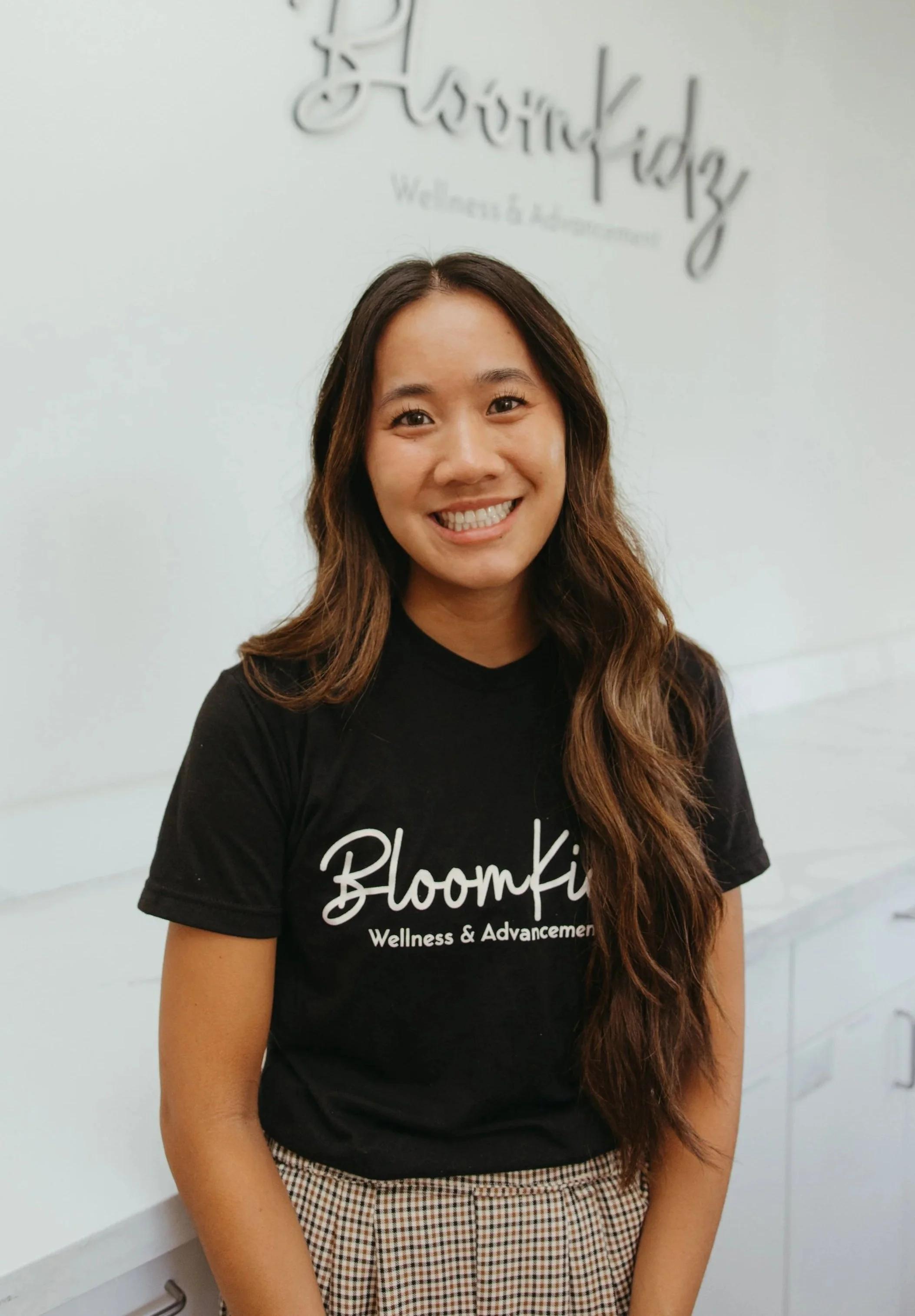 A smiling woman with long brown hair in a checkered skirt and a black shirt with white text reading 'BloomKidz Wellness & Advancement,' standing in front of a white wall with a sign that says the same