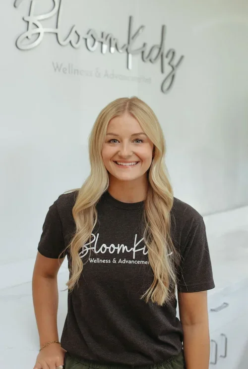 A smiling woman with long blonde hair standing indoors in front of a white wall with the words "Bloomkidz Wellness & Advancement" and "BloomKidz" written on it. She is wearing a black T-shirt with the same 'Bloomkidz' logo and text.