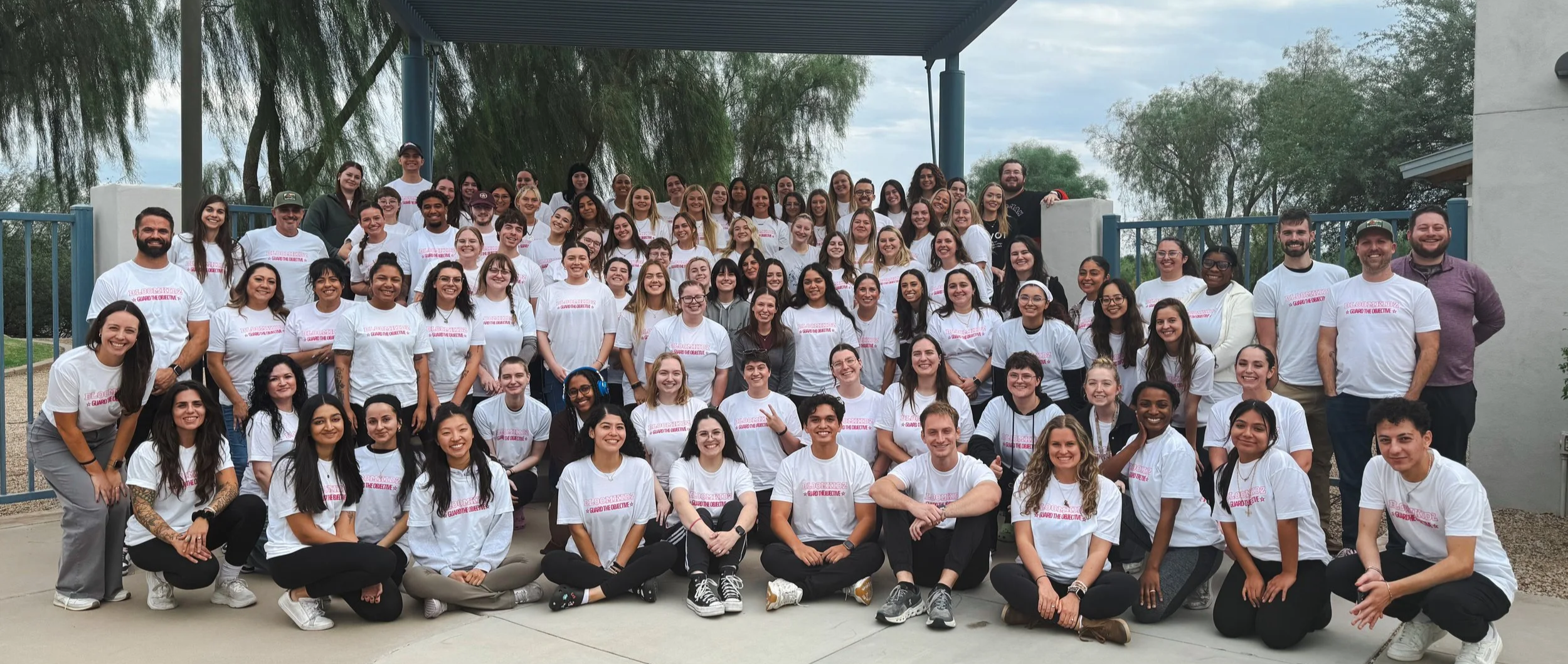 Large group of people wearing matching white T-shirts posing outdoors beneath a covered patio.