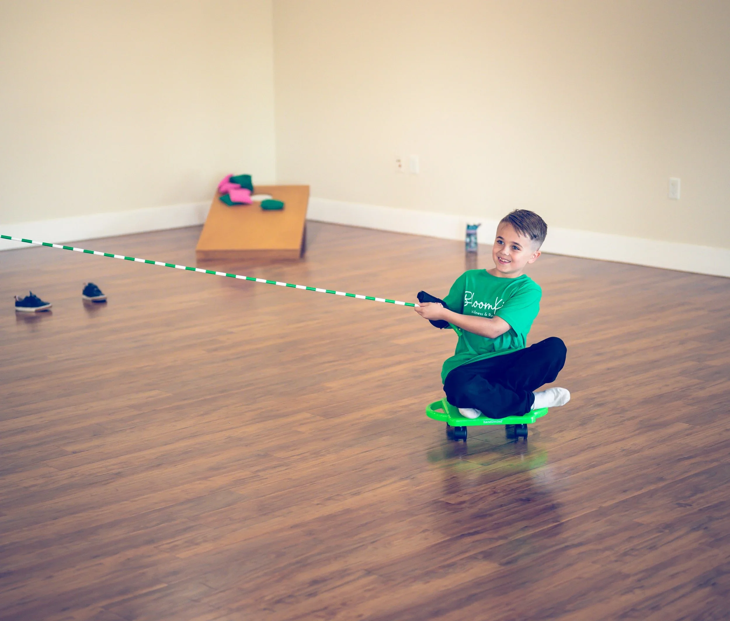 A young boy with a big smile sitting on a green ride-on toy in a room with hardwood floors and beige walls, holding a string connected to a pair of sneakers on the floor.