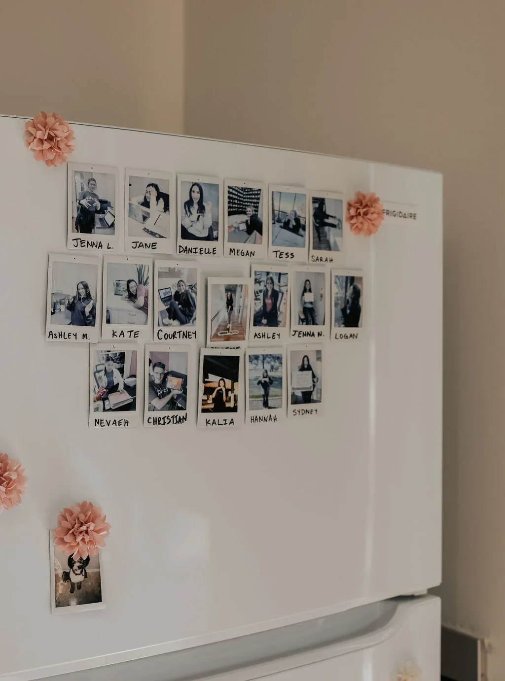 A white refrigerator door decorated with black-and-white Polaroid photos of employees, each labeled with their names. The photos are arranged in a grid, with some photo corners decorated with pink paper flowers.