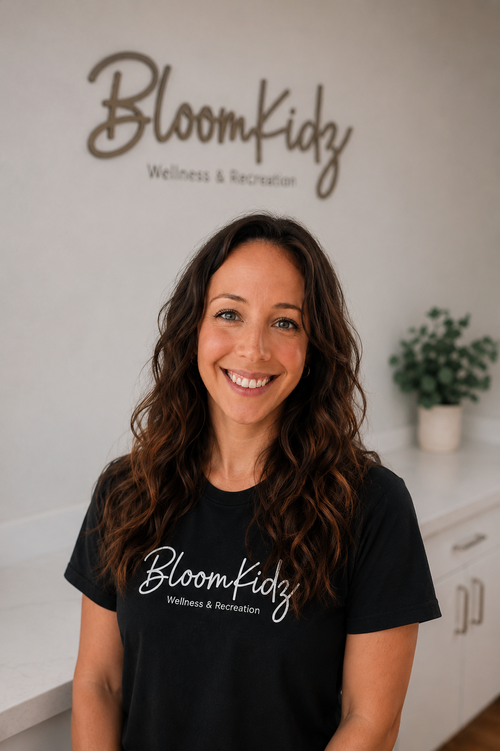 Woman with long curly brown hair smiling in front of a wall with Bloom Kidz Wellness & Recreation sign, wearing a black T-shirt with same logo.
