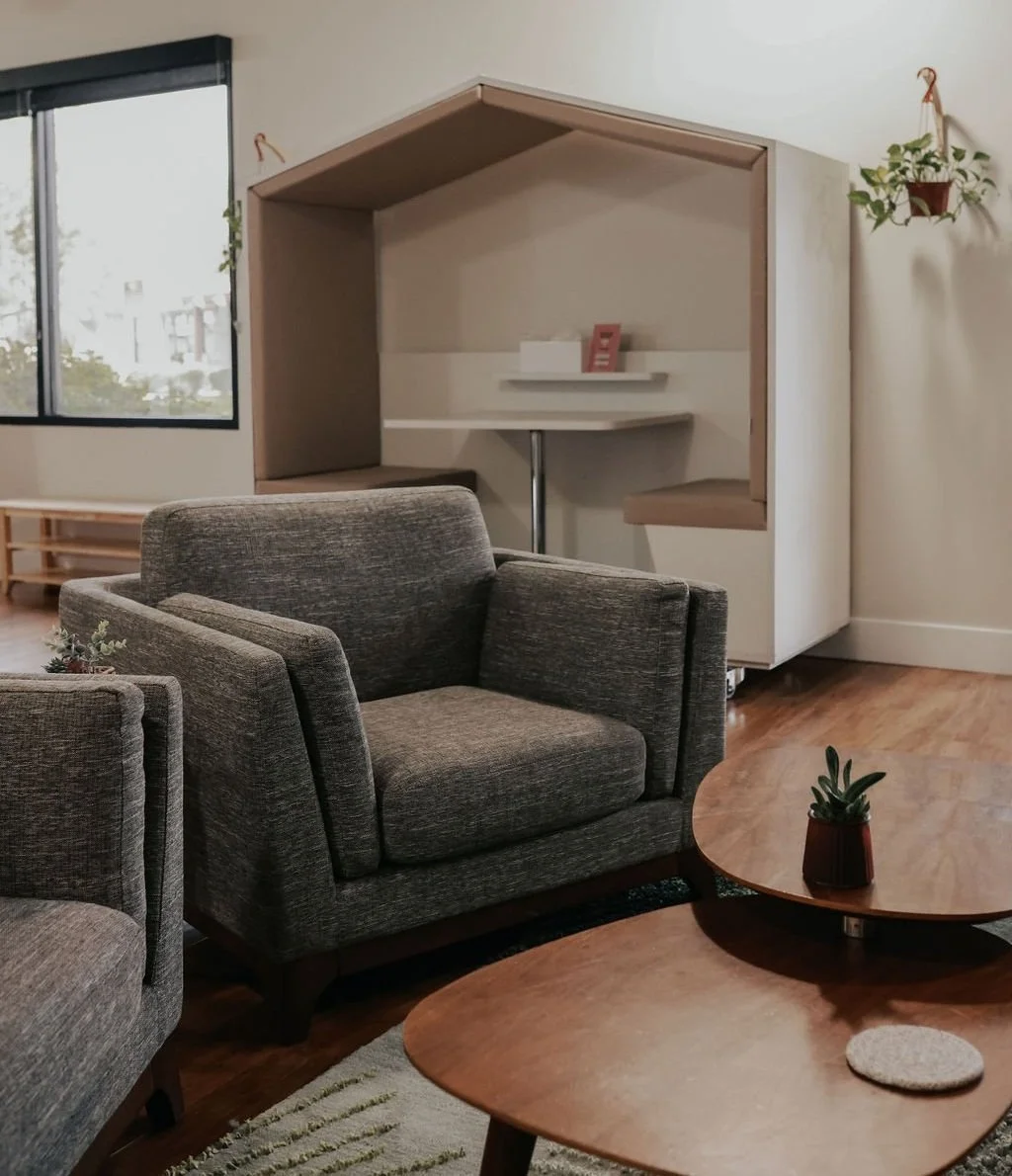 A cozy room with a gray armchair, a wooden coffee table with a small potted plant, and a beige booth with a table inside, situated in front of a window.