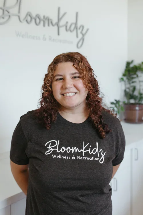A woman with curly red hair smiling, wearing a black T-shirt with 'Bloomkidz Wellness & Advancement' printed on it, standing indoors with a white wall and potted plant in the background.
