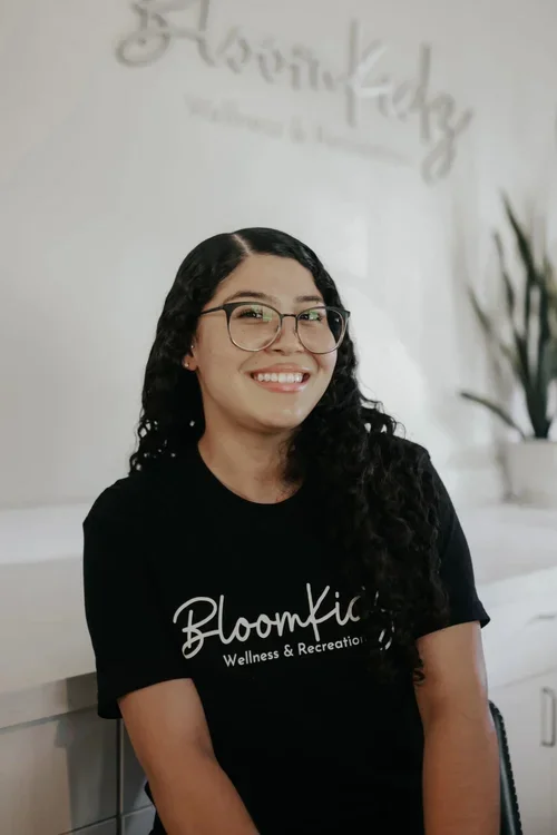 Woman with glasses and long curly hair smiling, wearing a black T-shirt that says BloomKidz Wellness & Recreation, in a wellness center with a plant in the background.