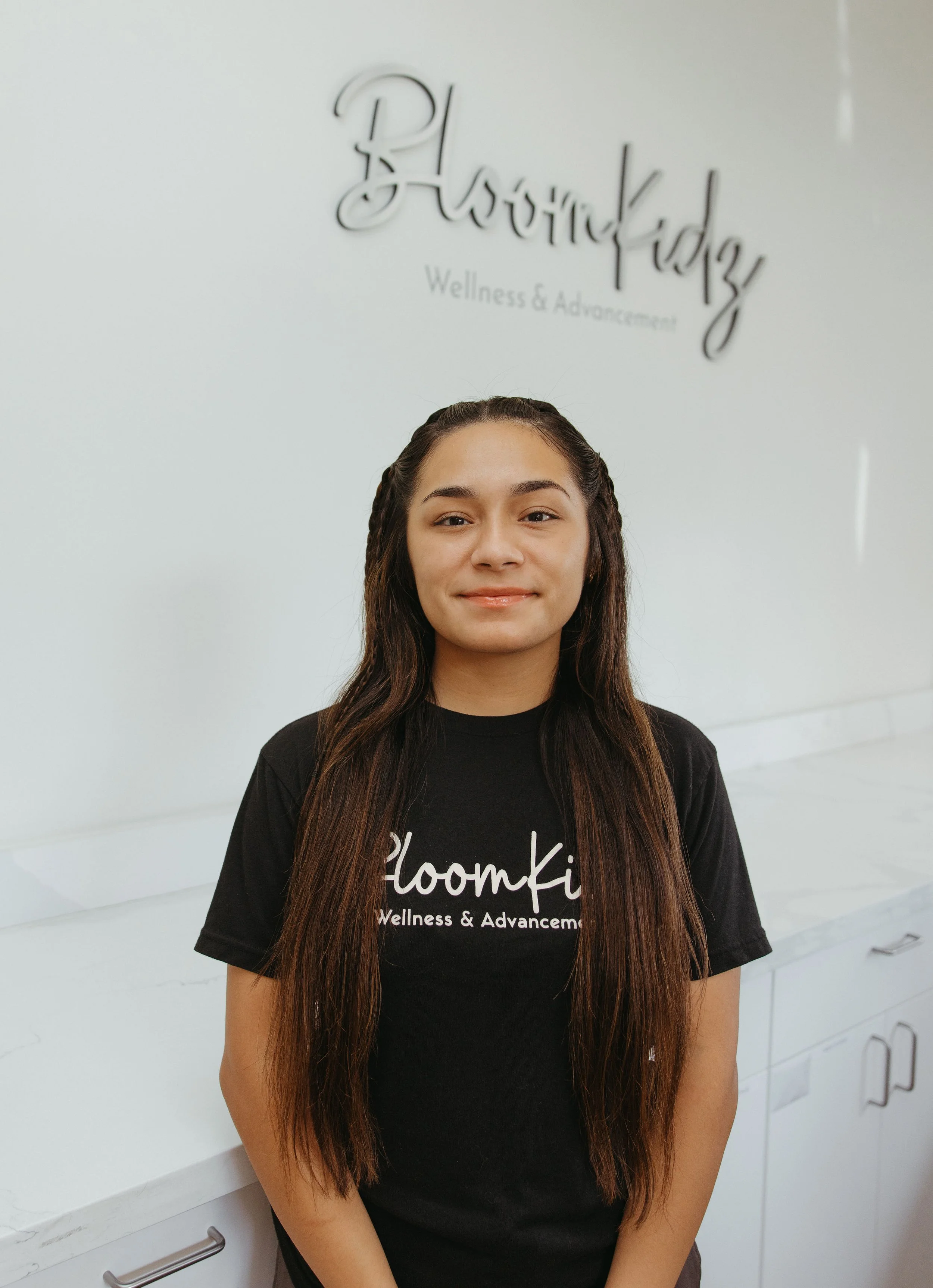 A young woman with long brown hair smiling, standing in front of a white wall with a sign that reads 'BloomKidz Wellness & Advancement'.