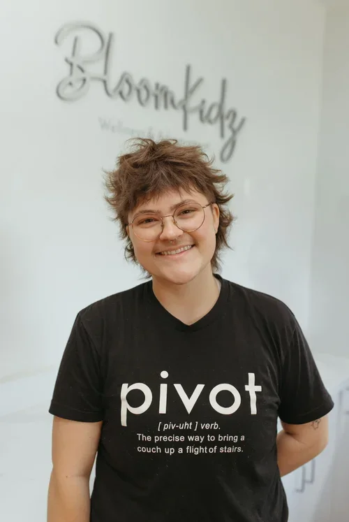 A smiling person with curly short hair, glasses, and a black pivot t-shirt standing indoors in front of a white wall with a sign that reads "BloomKidz".