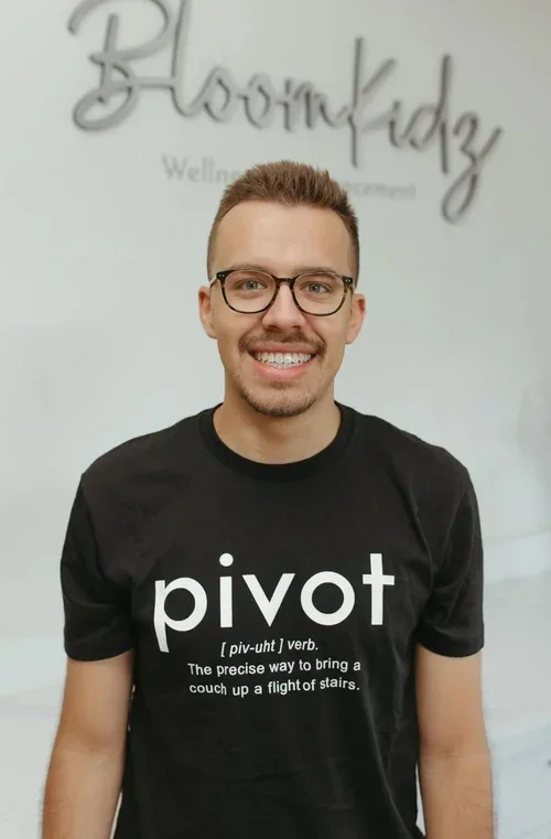 A smiling man with glasses and a beard wearing a black t-shirt with the word 'pivot' and a definition printed on it, standing indoors with a blurred background and decorative wall art.