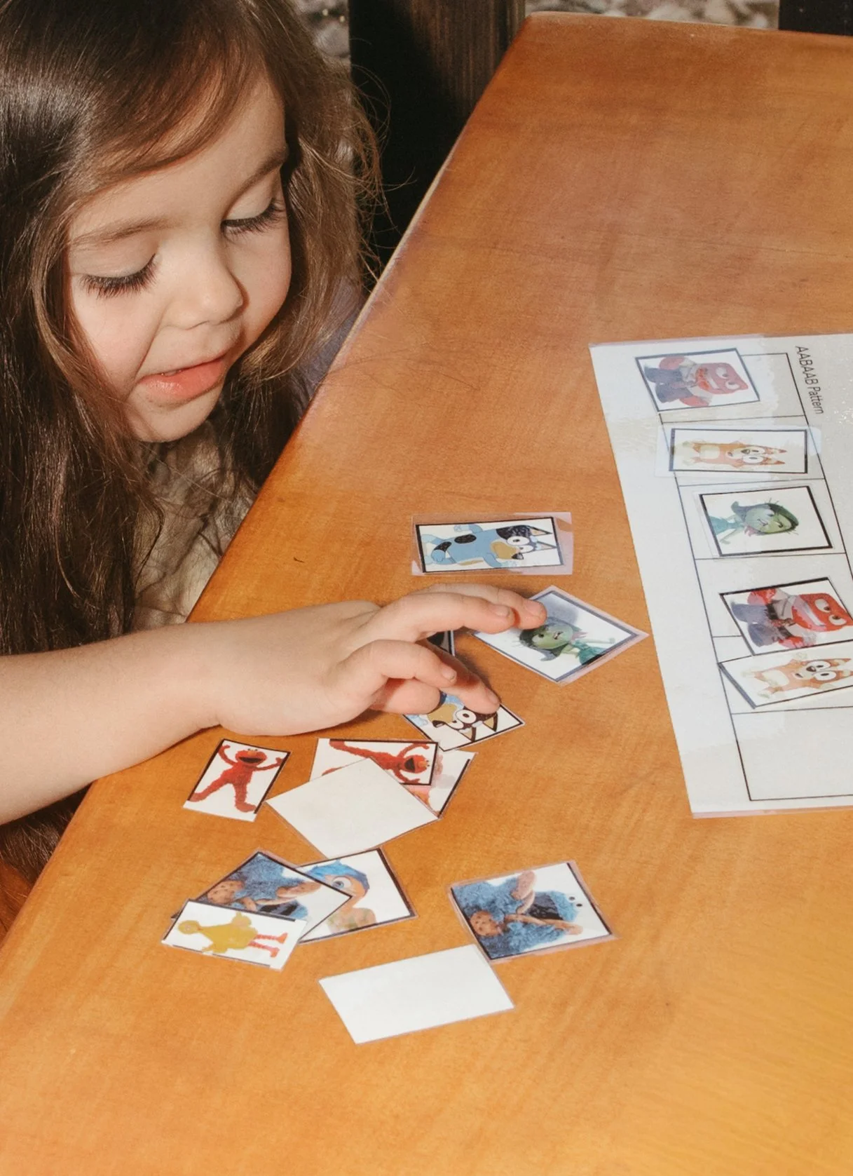 A young girl with long brown hair playing a matching game with Disney character cards on a wooden table. She has a gentle expression and is focused on the game. There are several cards face up and face down, and a game sheet with images of Disney characters in the background.
