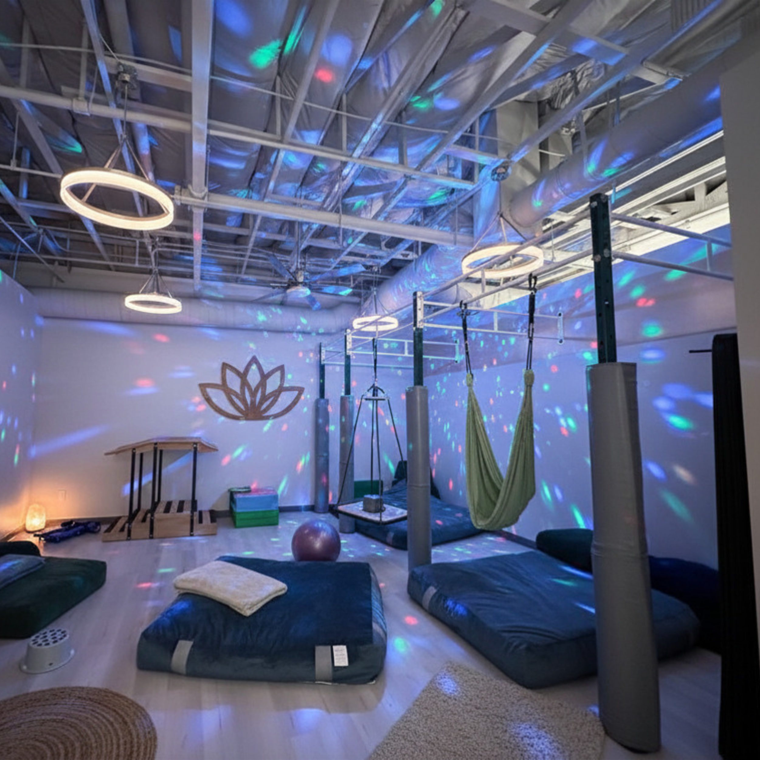 Indoor gym with metallic ceiling beams, colorful disco lights, ring-shaped lights, fitness equipment, mats, and hanging ropes.