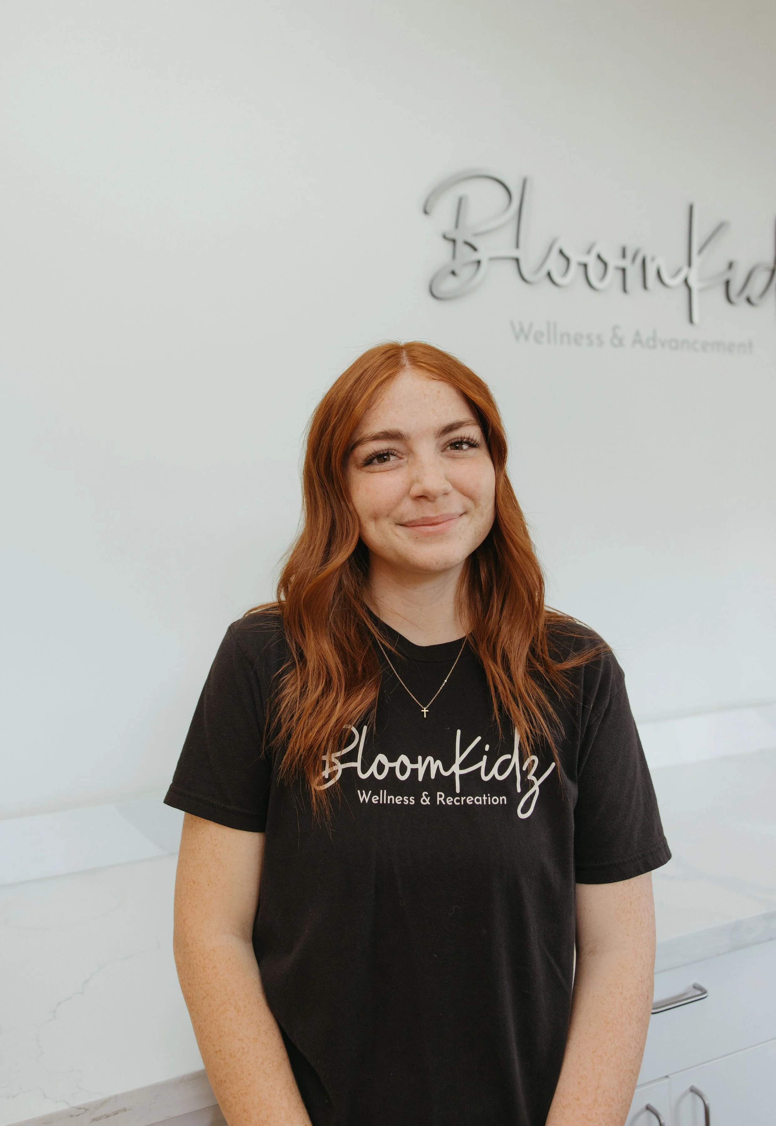 Young woman with red hair wearing a black t-shirt with white lettering and a cross necklace, standing in front of a white wall with a sign that reads 'Bloomkidz Wellness & Advancement'.