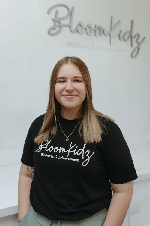 A young woman standing in front of a light-colored wall with the Bloomkidz logo and slogan, wearing a black T-shirt with the Bloomkidz logo and slogan, and gold necklace.