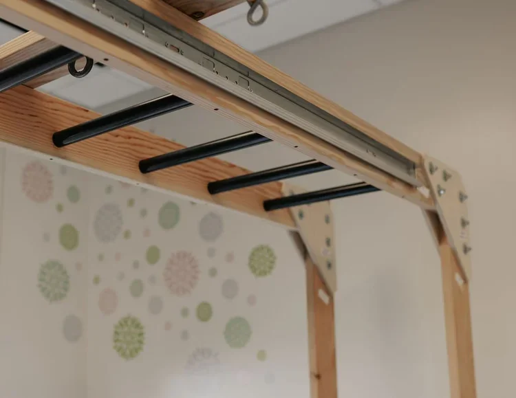 Part of a wooden monkey bar set with metal slats visible against a wall with decorative polka dot patterns.