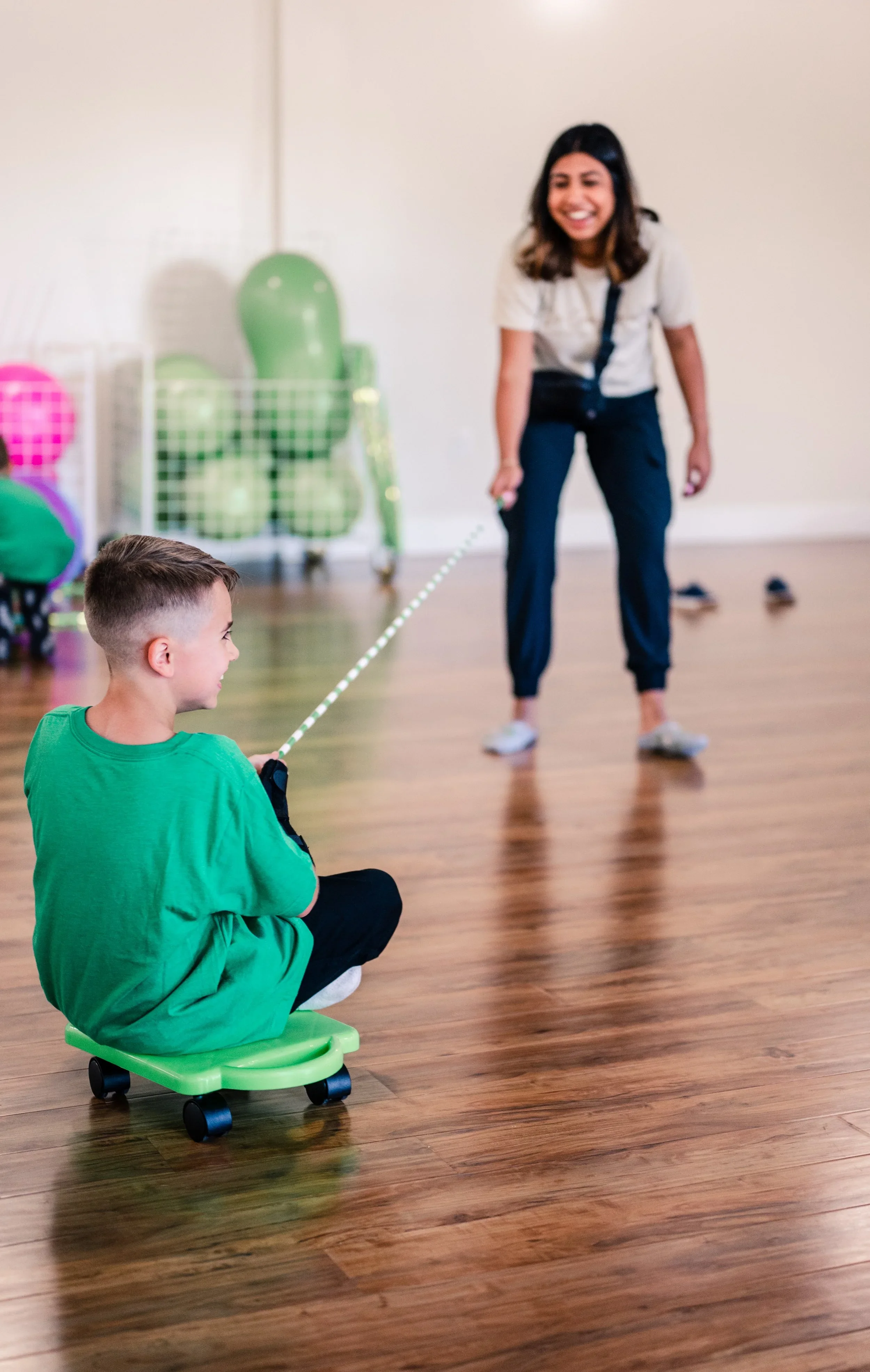 A young boy sitting on a small green ride-on toy, holding a one end of a rope, and smiling at a woman pulling him, holding the other end of the rope in a room with wooden floors and colorful exercise balls in the background.