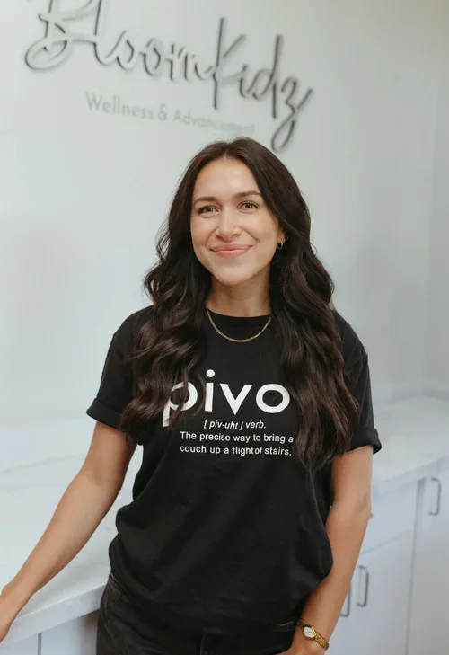 A woman with long, wavy dark hair smiling in front of a light-colored wall with a logo that reads 'BloomKidz Wellness & Advancement'. She is wearing a black t-shirt with the word 'pivot' and its definition on it.