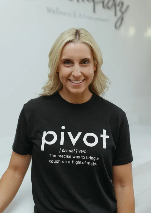 Woman with blonde hair smiling, wearing a black t-shirt with the word 'pivot' and its definition printed on it, standing against a light background.