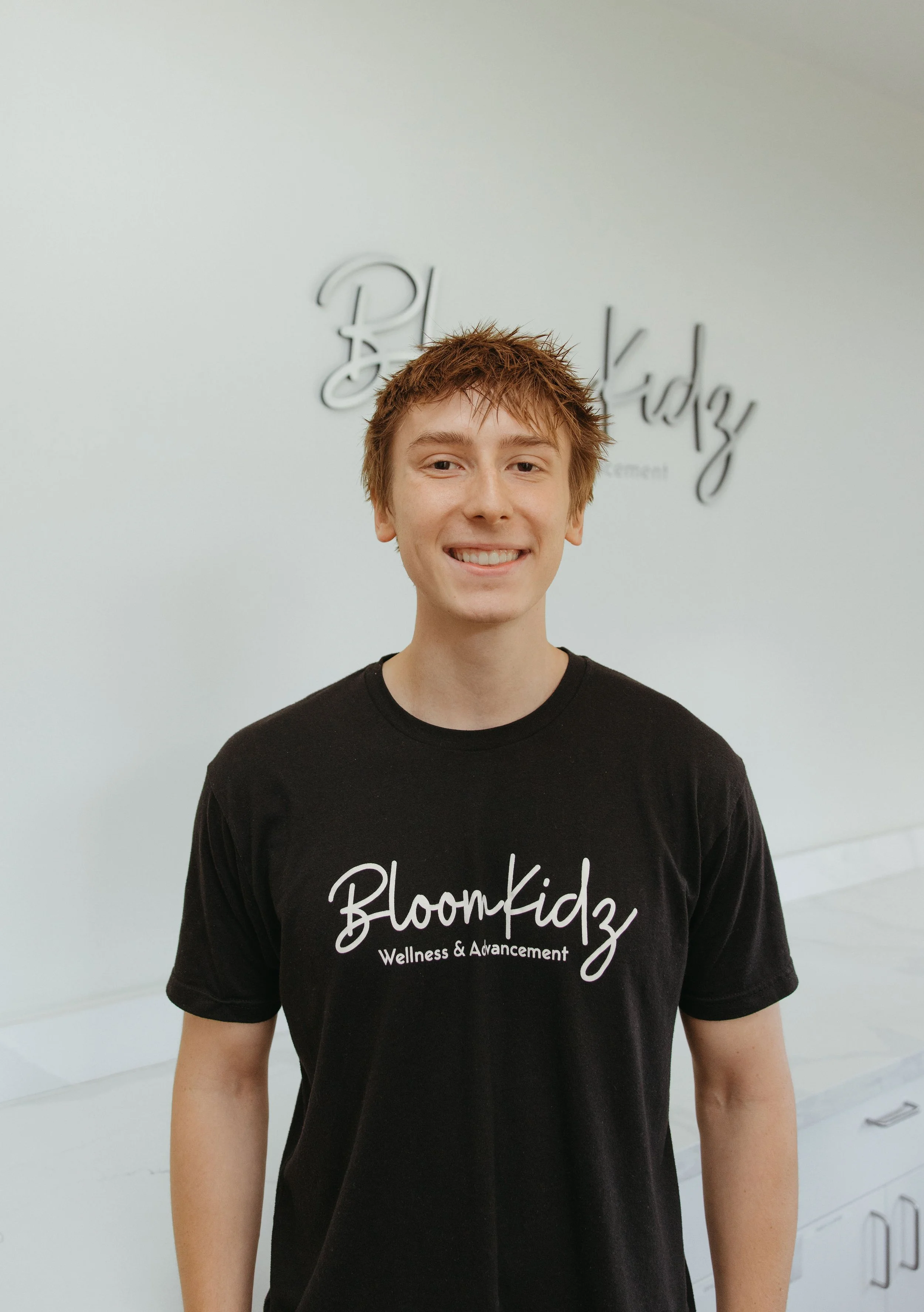 Young person with short, messy red hair smiling, wearing a black t-shirt with white text that reads 'BloomKidz Wellness & Advancement,' standing in front of a light-colored wall with the words 'BloomKidz' written in a cursive font.