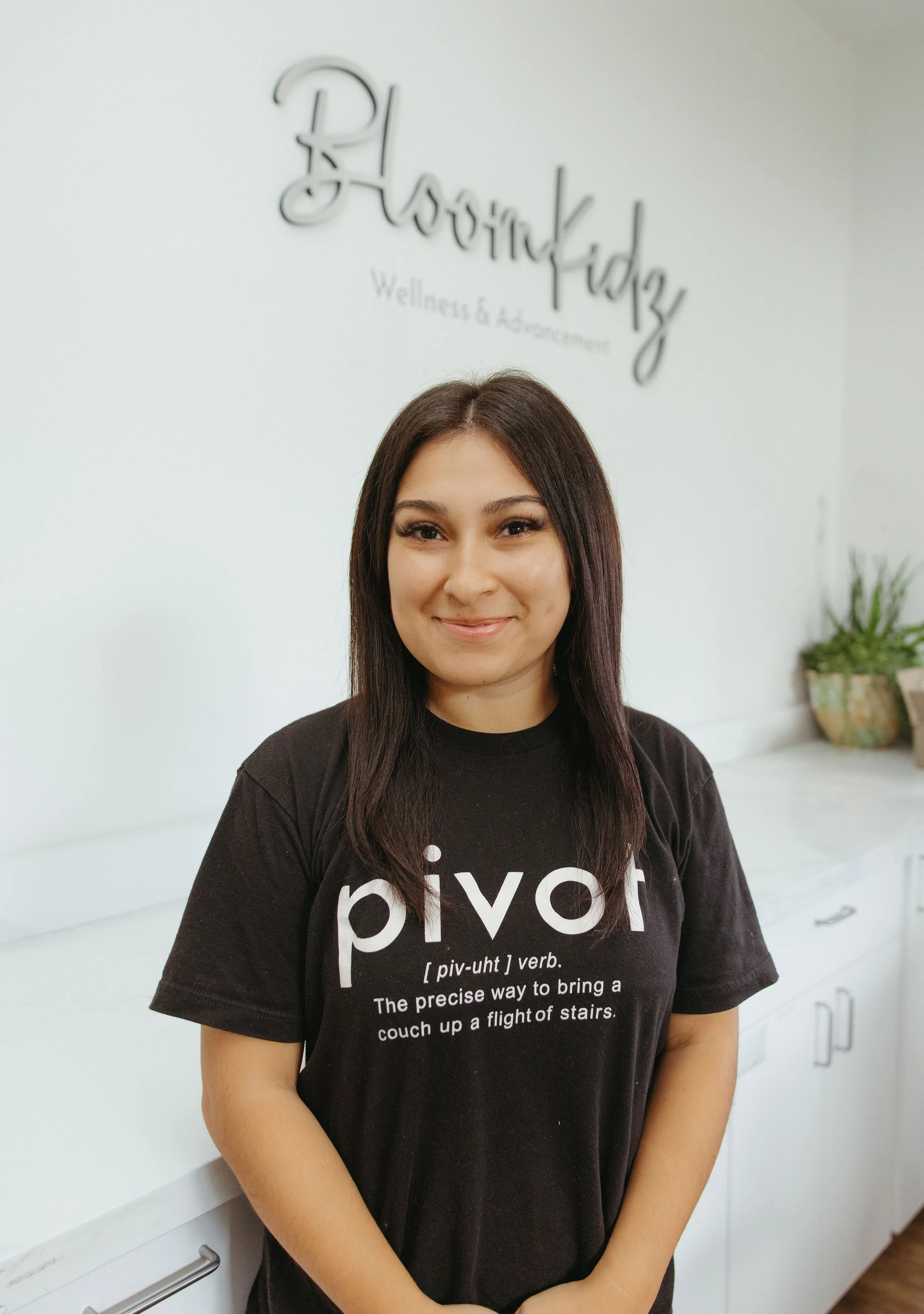 Smiling woman wearing a black “pivot” T-shirt standing in a bright kitchen