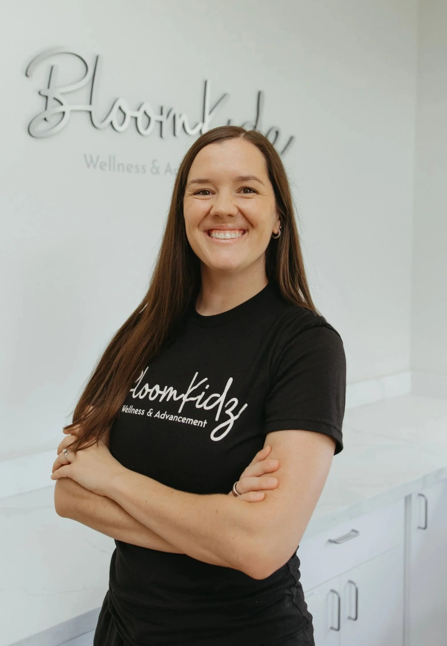 A smiling woman wearing a black t-shirt with Bloomkidz Wellness & Recreation logo, standing in a wellness center with a sign and a plant in the background.