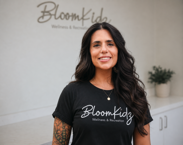 A woman with long dark hair smiling, wearing a black Bloomkidz Wellness & Recreation t-shirt, standing indoors with a white wall and potted plant in the background.